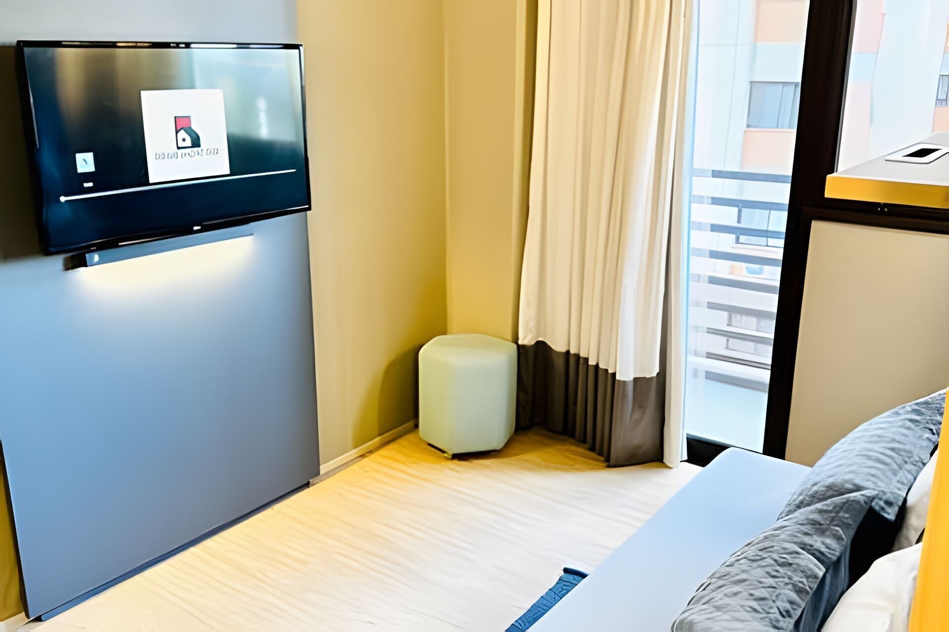 Habitación de hotel con televisor de pantalla plana montado en la pared, un cubo de basura pequeño, una cama con almohada a rayas azules y una ventana con cortinas.