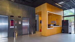 A modern elevator with a digital display and emergency buttons, a reception desk with a wooden finish, and a large window providing a view of the outdoors. - Prime - Flats - Alpha Stay (Barueri)