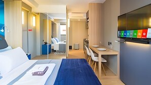 A hotel room with a bed, a desk, a chair, a television, and a view of the hallway. - Prime - Flats - Alpha Stay (Barueri)