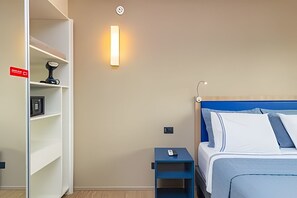Habitación de hotel con cama, mesita de noche, estante y lámpara de pared.