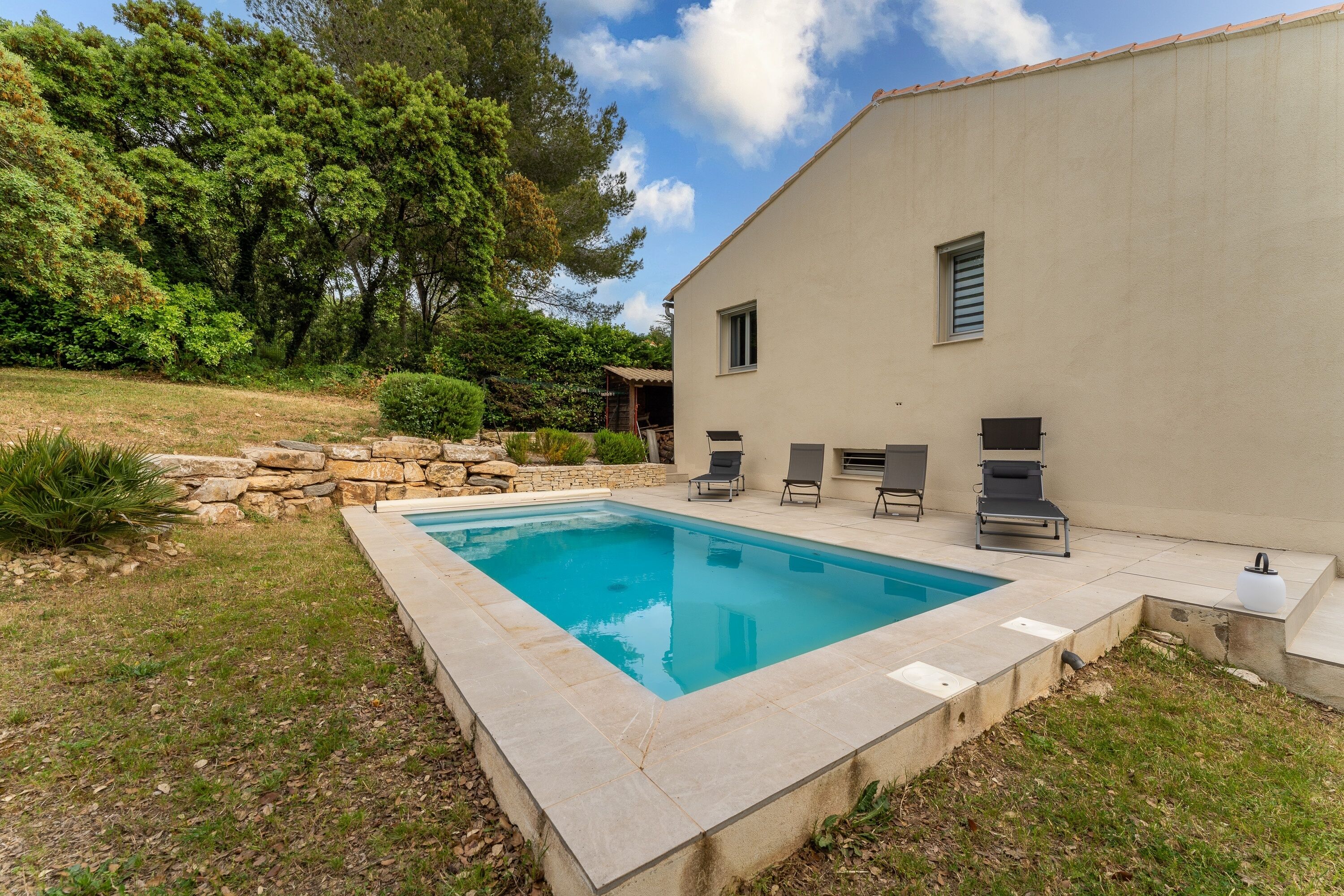 Una piscina con borde de piedra, un patio con sillones de descanso y un edificio de fachada color beige.