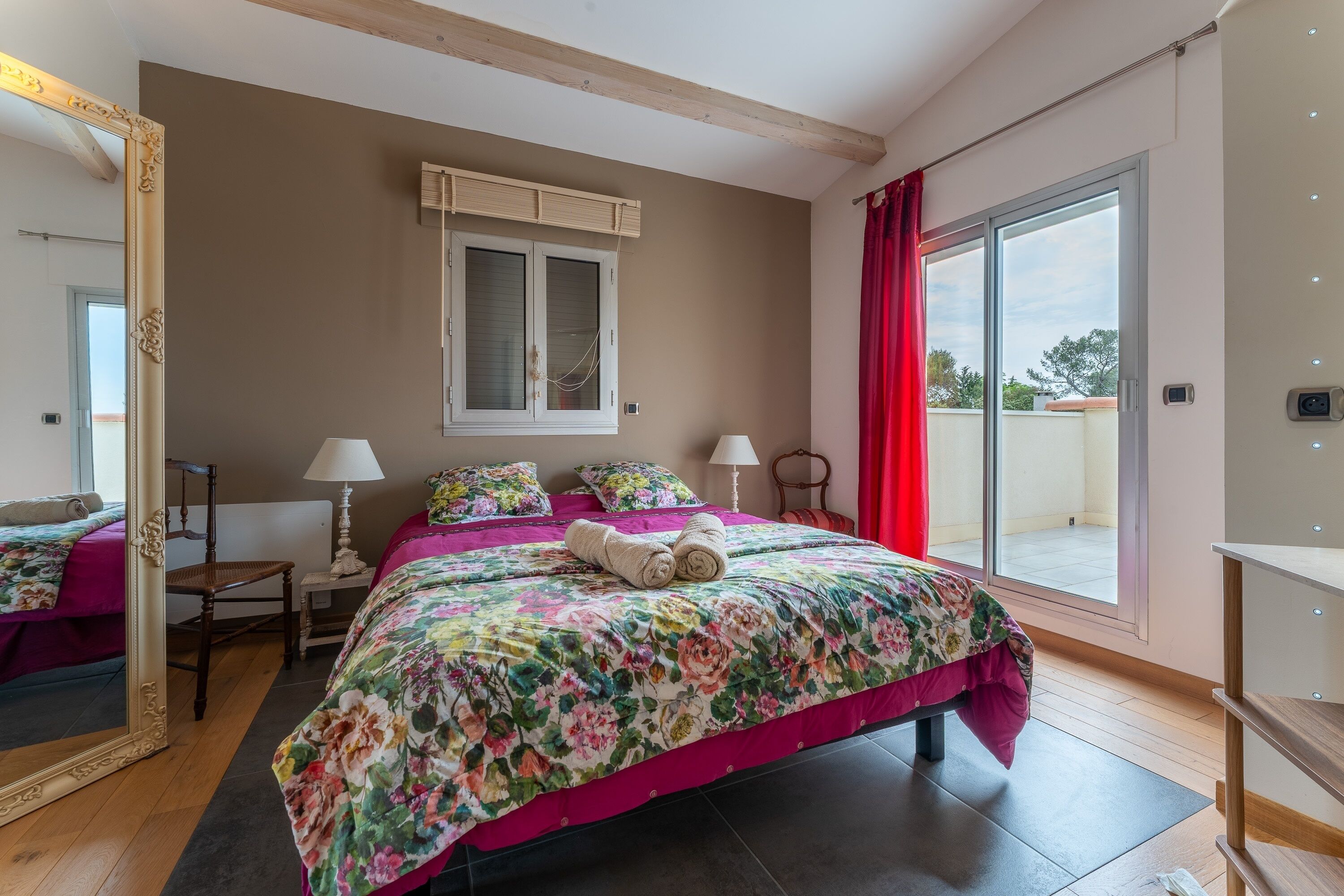 Un dormitorio con cama, ventana con cortinas, espejo y suelo de madera.