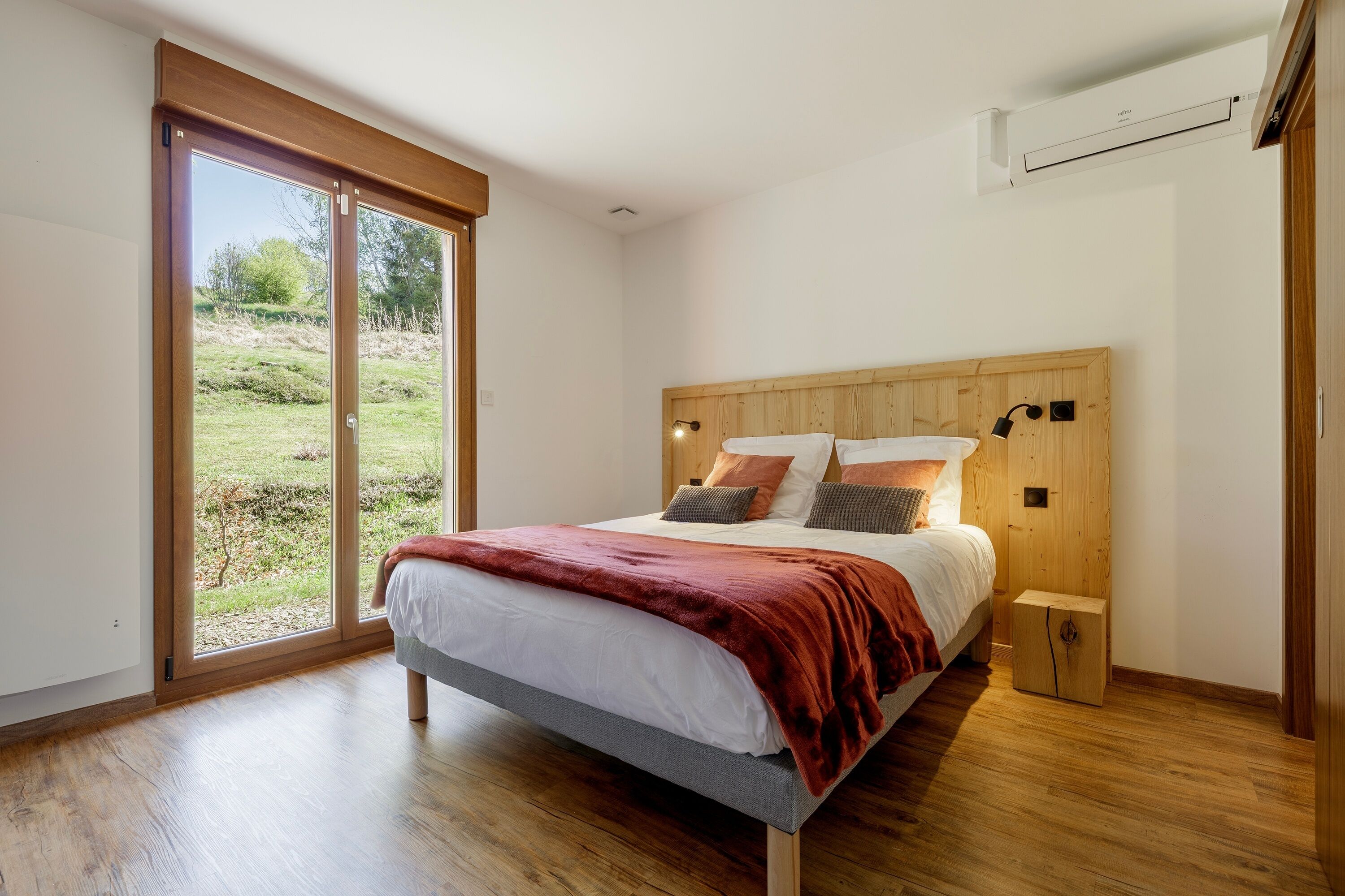 Un dormitorio con cama, mesita de noche, cabecera de madera, ventana y unidad de aire acondicionado.