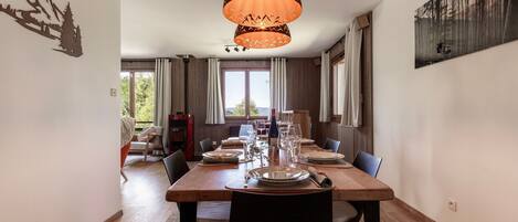 Comedor con mesa de madera dispuesta para cuatro, una lámpara colgante y vista al exterior a través de amplios ventanales.
