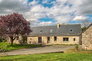 Une maison en pierre avec un toit en pente, une allée pavée et un arbre aux fleurs roses.