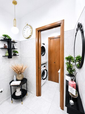 A modern bathroom with a wooden door, a washing machine, a clock, and a shelf with plants. - Elegant 3BR Apartment with Ensuite Baths in Central Naples (Napoli)