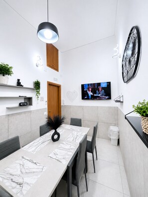 A modern dining area with a marble table, black chairs, a black pendant light, a television on the wall, and a large clock. - Elegant 3BR Apartment with Ensuite Baths in Central Naples (Napoli)