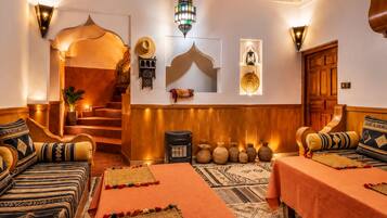 A traditional Moroccan interior with a staircase, patterned sofas, a central rug, and decorative wall sconces.