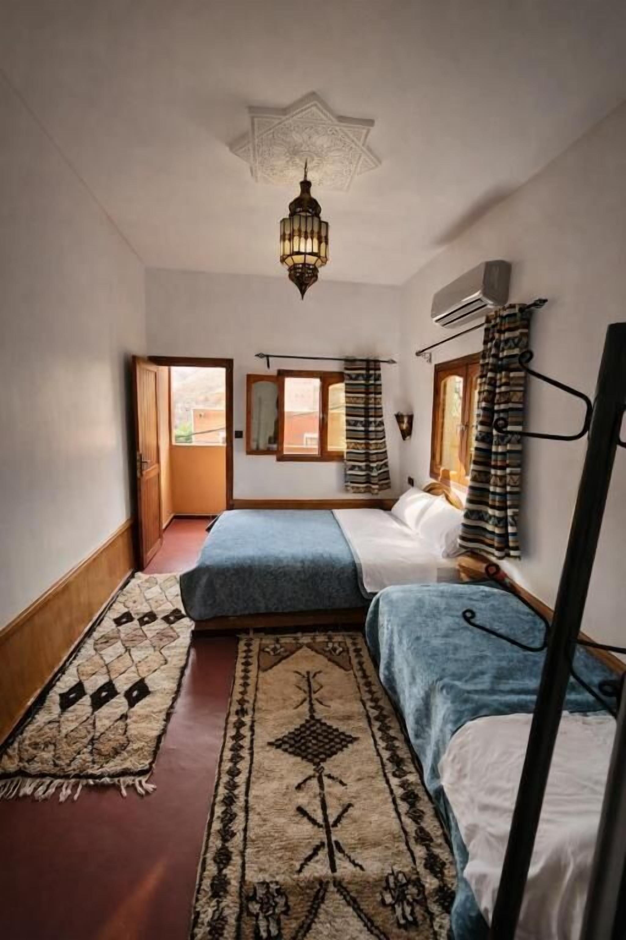 Une chambre avec deux lits, un tapis à motifs, un luminaire suspendu et une fenêtre avec des rideaux.
