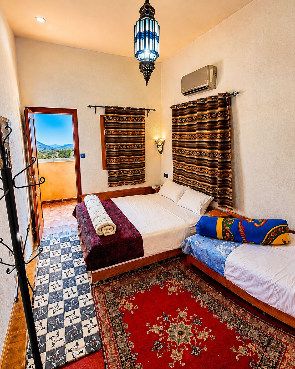 A hotel room with a bed, a red patterned rug, a wooden floor, a wall-mounted air conditioner, and a view of the outdoors through a door.