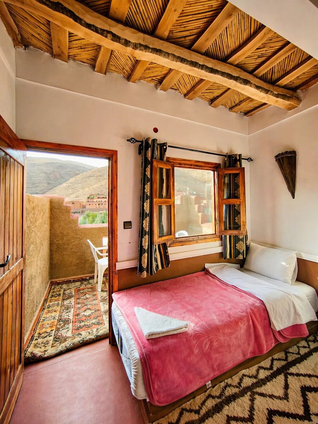 Ein Schlafzimmer mit Holzbalkendecke, einem Fenster mit gemusterten Vorhängen, einem Bett mit rosa Decke und Blick auf eine gebirgige Landschaft.