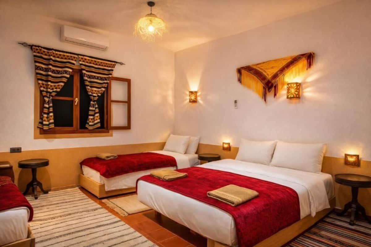 A hotel room with two beds, a window with curtains, a wall-mounted light, and a small table.