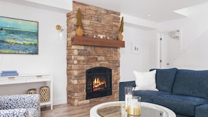 A living room with a stone fireplace, a blue sofa, a glass coffee table, and a painting on the wall. - Private lakefront retreat in Muskoka w/firepit, hot tub, private dock, kayaks (Huntsville)