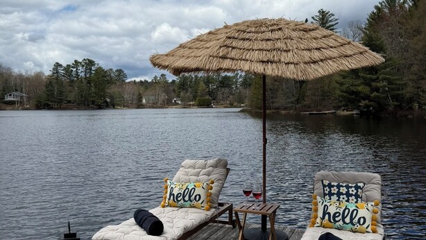A wooden deck with two lounge chairs, a table, a lantern, and a thatched umbrella. - Private lakefront retreat in Muskoka w/firepit, hot tub, private dock, kayaks (Huntsville)