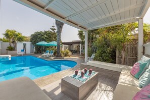 Ein Poolbereich mit überdachter Terrasse, Liegestühlen und einer Sitzecke mit Kissen.