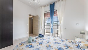 A bedroom with a bed, a window with curtains, a door, and a nightstand with a lamp. - Villa Cotriero Crystal (Gallipoli)