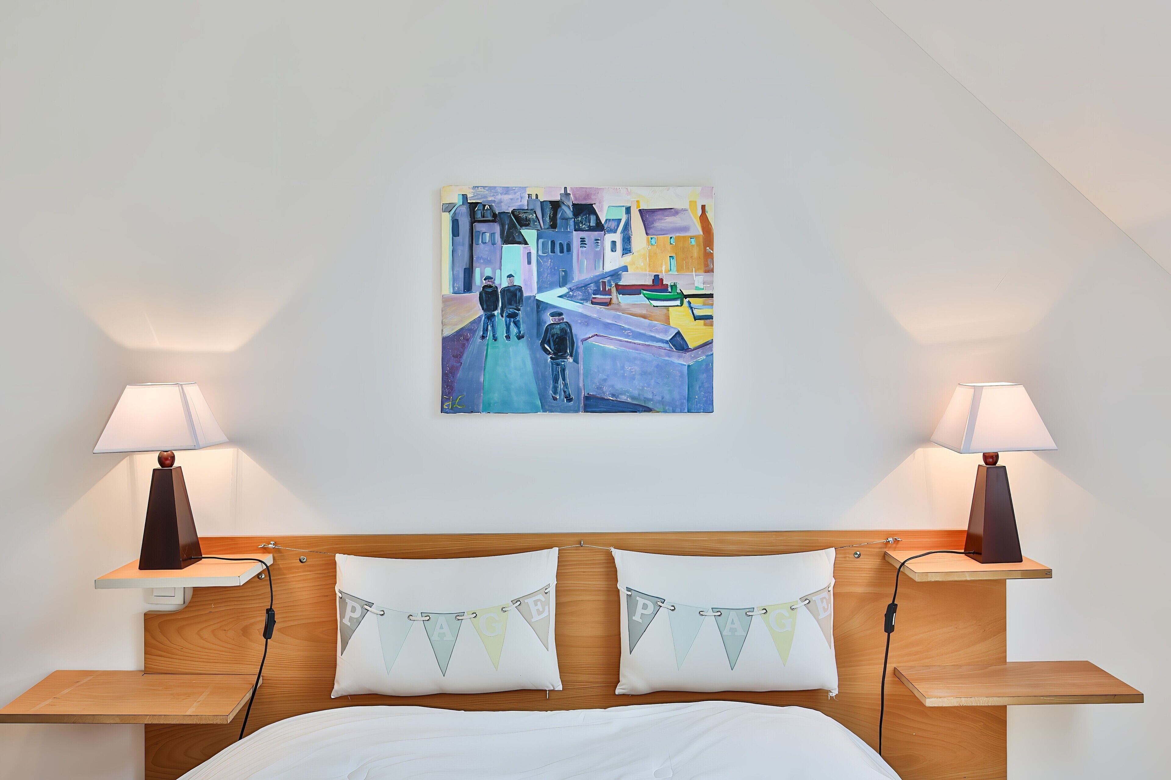Ein Hotelzimmer mit einem Bett, zwei Nachttischen mit Lampen, einem Wandbild und einem Kissen mit dem Buchstaben 'P'.