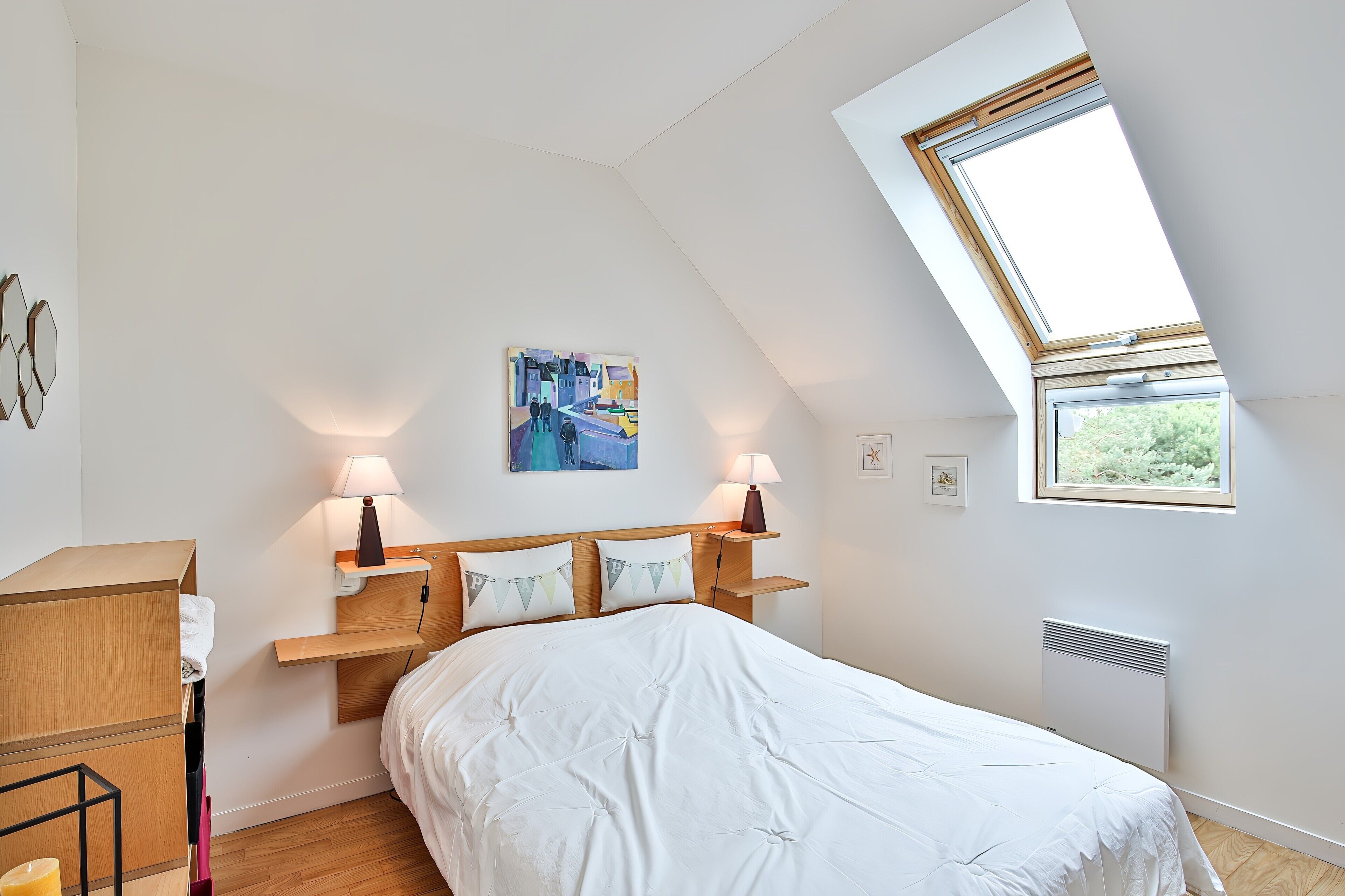 Une chambre avec un plafond en pente, un puits de lumière, un lit avec une literie blanche, deux lampes de chevet, un tableau au mur et un radiateur.