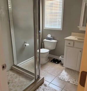 Une salle de bain avec une porte de douche en verre, des toilettes, un lavabo et une fenêtre avec des stores.