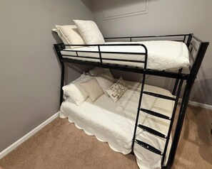 Un lit superposé avec un matelas et des oreillers blancs, une échelle et un sol recouvert d’un tapis beige.