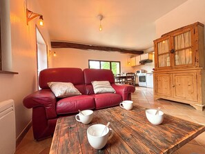 Ein rotes Ledersofa mit Kissen, ein Holzesstisch mit weißen Schüsseln und Löffeln, ein Holzschrank und eine Küche im Hintergrund.