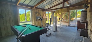 Ein Billardtisch, Fitnessgeräte und ein Punktsack in einem Raum mit Holzwänden und großen Fenstern.