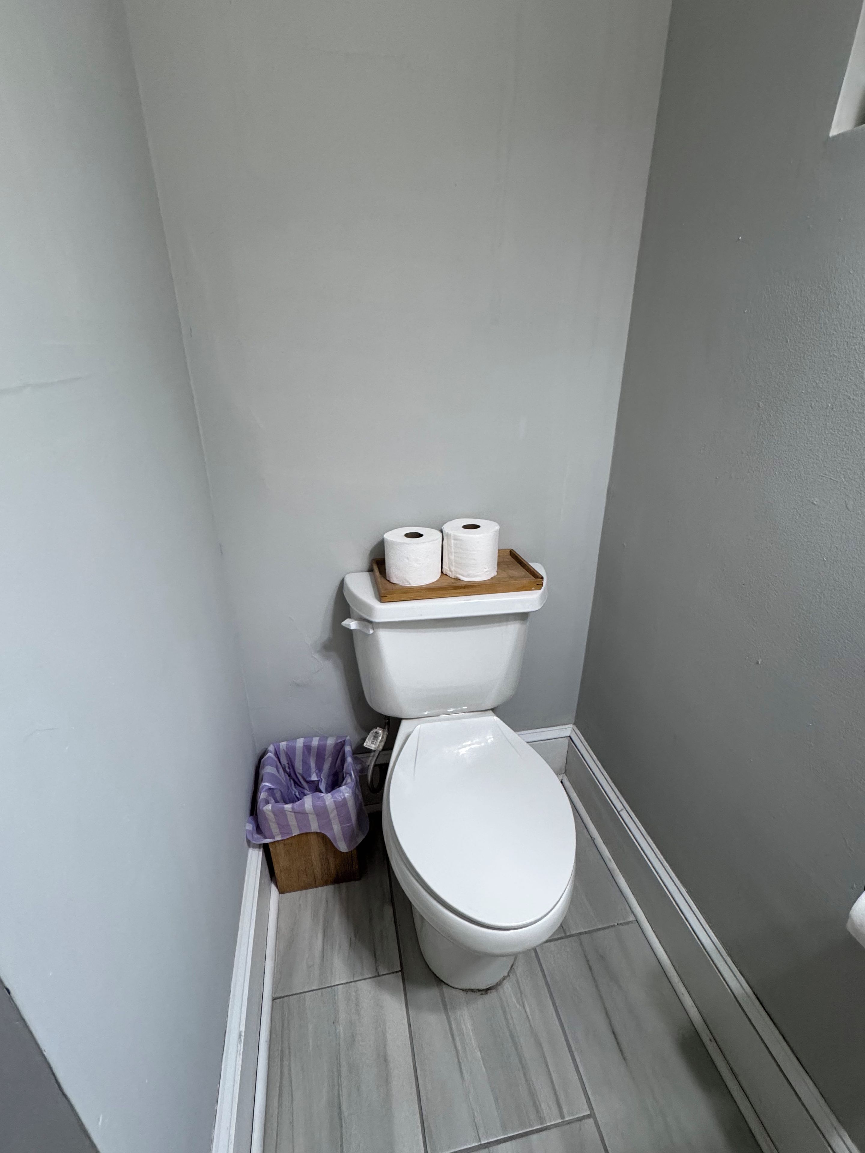 A small bathroom with a toilet, a wooden toilet paper holder, and a trash bin.