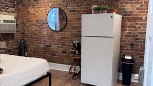 A room with a brick wall, a bed with a white blanket, a white refrigerator, a round mirror, a small table with a coffee maker, and a wooden floor. - Jungle House Studio Suites – Short North & Convention Center (Columbus)
