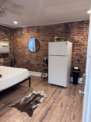 A room with a brick wall, a bed with a white blanket, a white refrigerator, a round mirror, a small table with a coffee maker, and a wooden floor.
