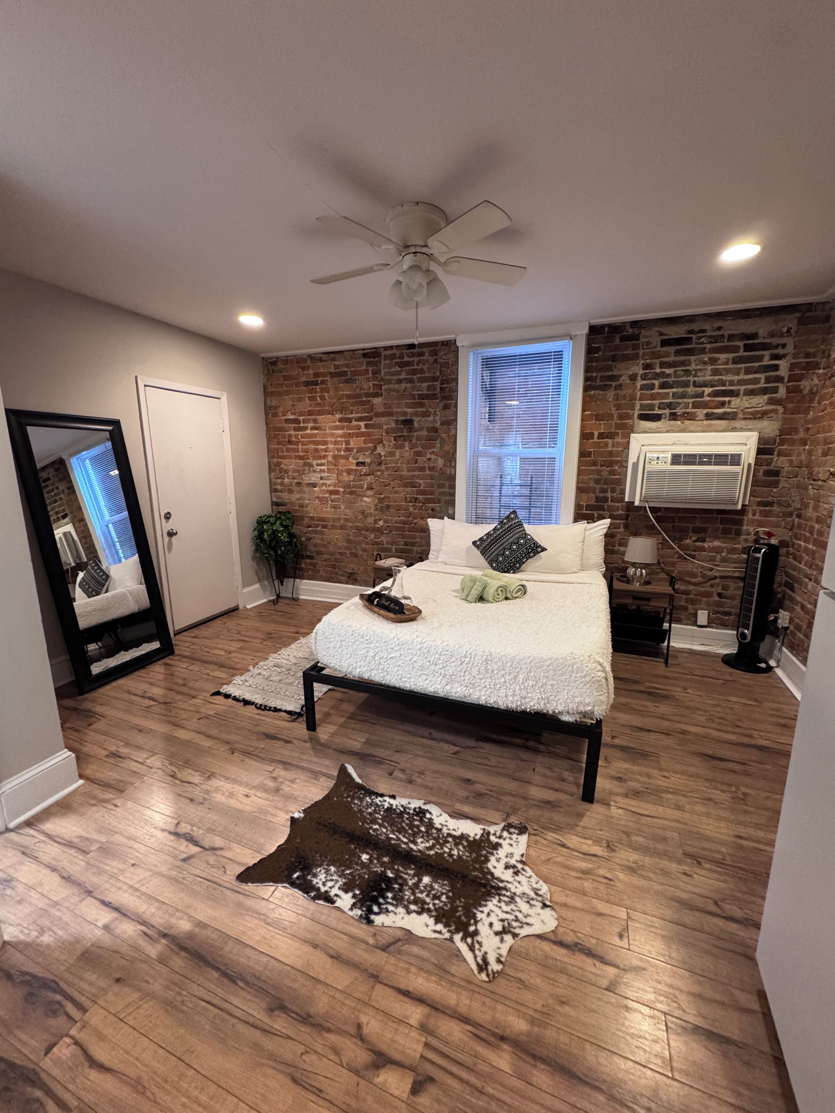 A bedroom with a bed, a ceiling fan, a brick wall, a window with blinds, and a wooden floor.