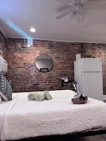 Una camera da letto con parete in mattoni, uno specchio tondo, un letto con biancheria bianca e un ventilatore a soffitto, oltre a un frigorifero bianco.