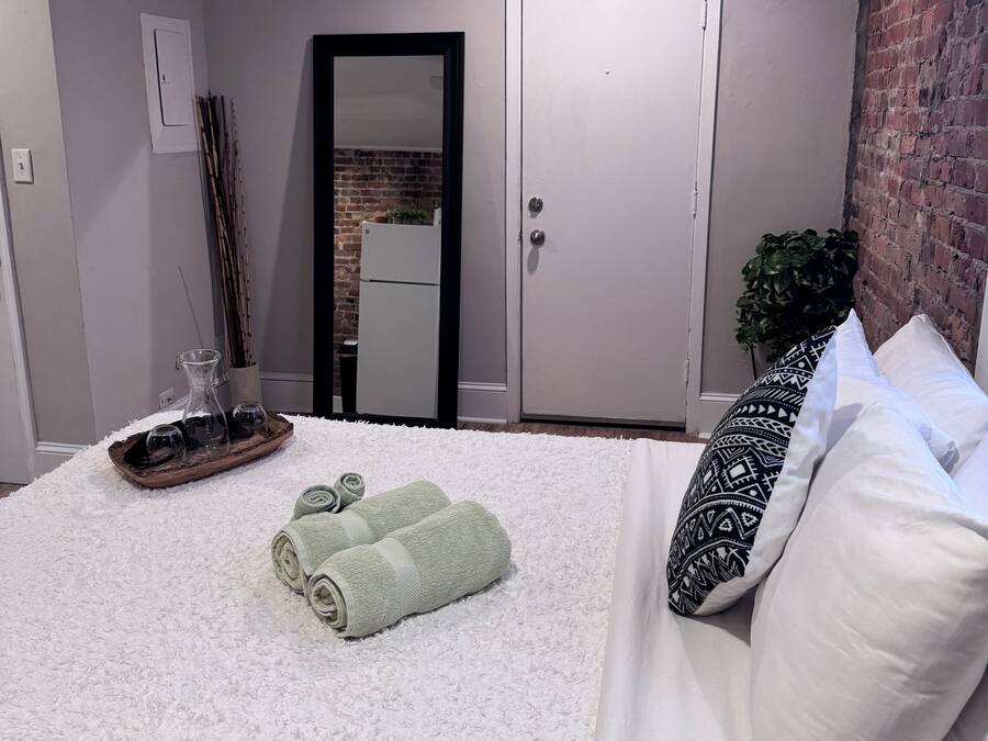 Quarto com cama, travesseiros, mesa de cabeceira e um copo sobre ela, e uma porta.