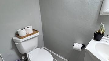 A bathroom with a toilet, a sink, a small shelf with a jar, and a trash bin.