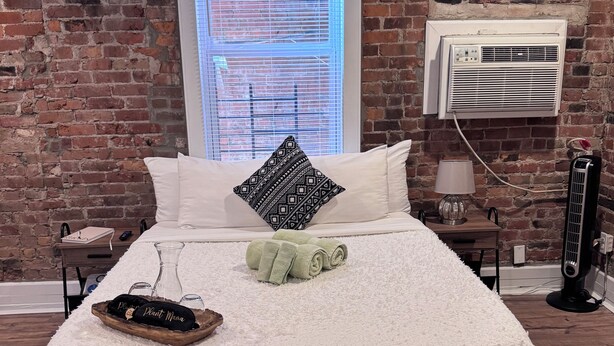 A bed with a white fluffy cover, a patterned pillow, and rolled towels. There is a small wooden tray with a plant and a bottle. A window with blinds is visible, and an air conditioning unit is mounted on the brick wall. - Jungle House Studio Suites – Short North & Convention Center (Columbus)