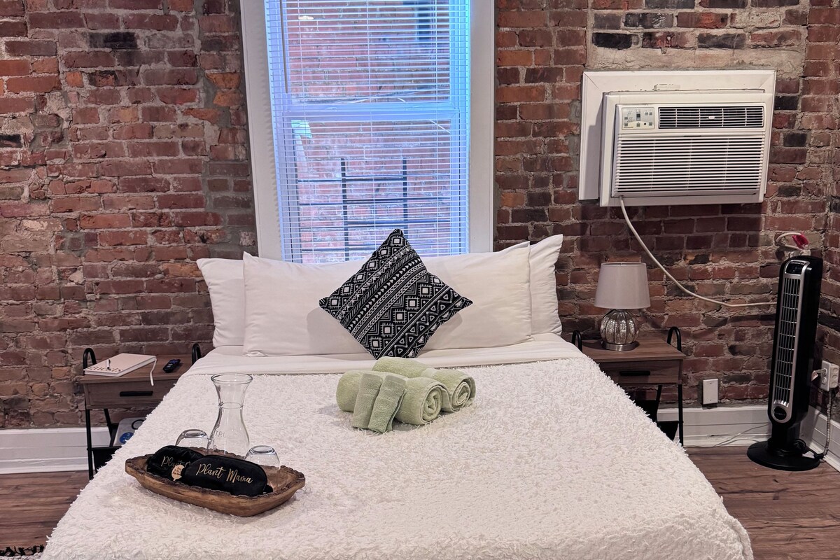 A bed with a white fluffy cover, a patterned pillow, and rolled towels. There is a small wooden tray with a plant and a bottle. A window with blinds is visible, and an air conditioning unit is mounted on the brick wall.