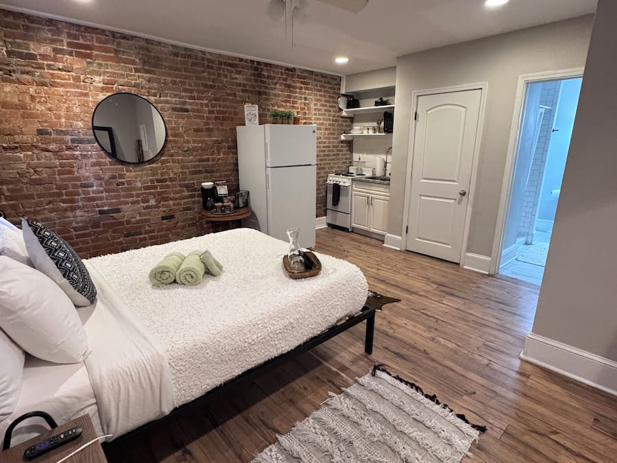 A bedroom with a bed, a round mirror, a ceiling fan, a brick wall, a kitchen area, and a wooden floor.