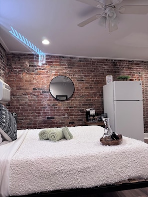 Un dormitorio con pared de ladrillo, un espejo redondo, una cama con ropa de cama blanca, un ventilador de techo y un refrigerador blanco.