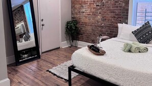 A bedroom with a brick wall, a ceiling fan, a bed with white bedding, a mirror, and a wooden floor. - Jungle House Studio Suites – Short North & Convention Center (Columbus)