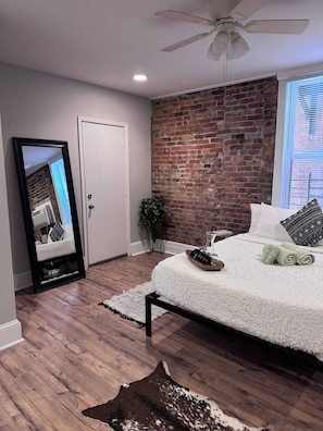 A bedroom with a brick wall, a ceiling fan, a bed with white bedding, a mirror, and a wooden floor.