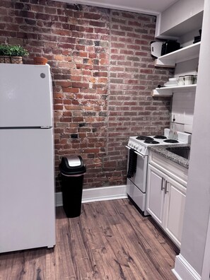 Una cocina compacta con pared de ladrillo, armarios blancos, un refrigerador de acero inoxidable, un microondas y una estufa a gas.
