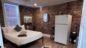 A bedroom with a bed, a ceiling fan, a brick wall, a window, a refrigerator, and a rug. - Jungle House Studio Suites – Short North & Convention Center (Columbus)