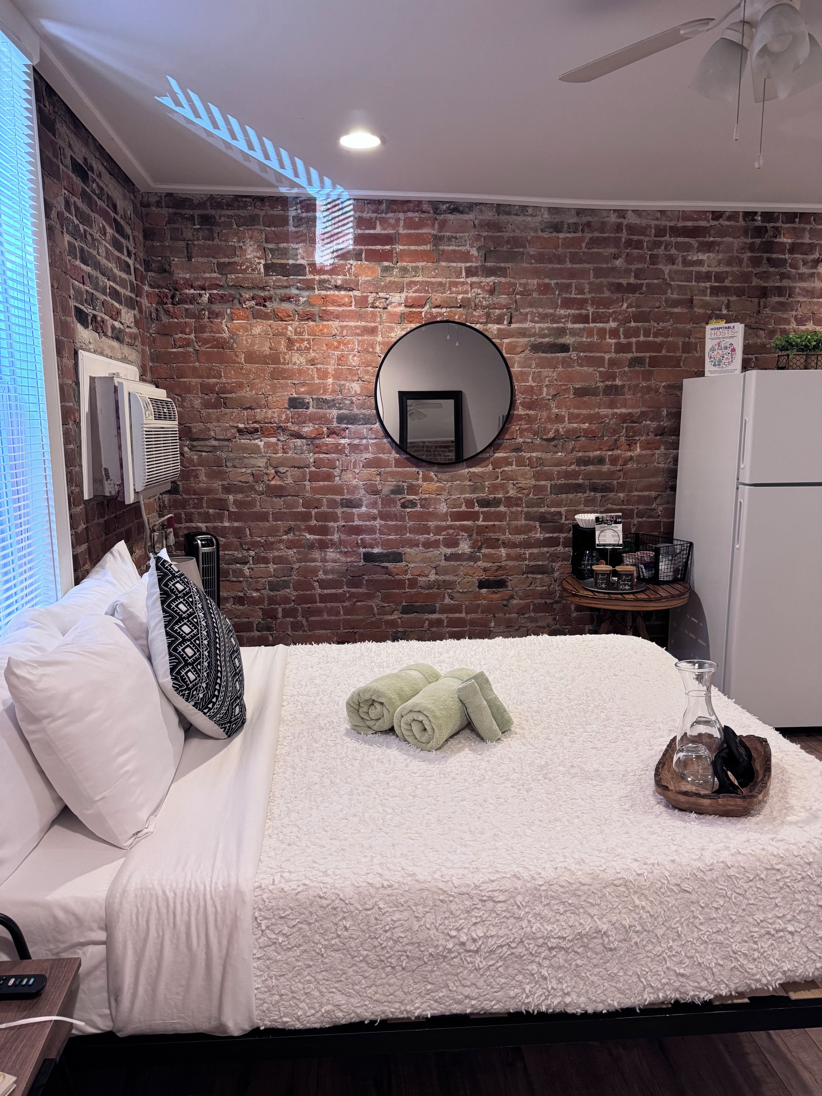 Habitación de hotel con pared de ladrillo, un espejo redondo, una cama con ropa de cama blanca, una alfombra blanca y una mesita con una planta.