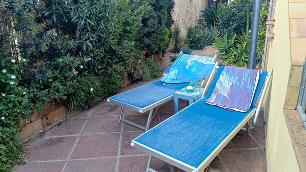 Twee ligbedden met blauwe kussens, een tafeltje met een schaal lekkernijen, en een tuin met diverse planten en bloemen.