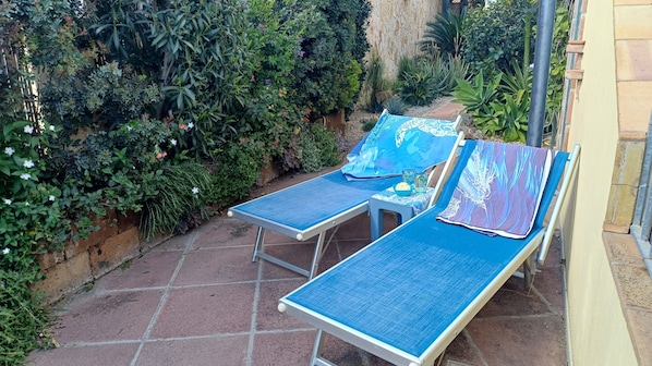 Duas espreguiçadeiras com almofadas azuis, uma mesa pequena com uma tigela de lanches, e um jardim com diversas plantas e flores.