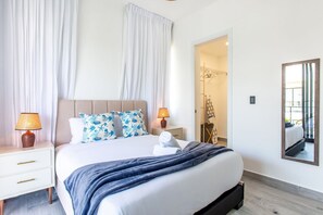 A hotel room with a bed, nightstands, a lamp, a mirror, and a door leading to a bathroom. - Modern 1BR, pools, gym, and artificial beach (Punta Cana)