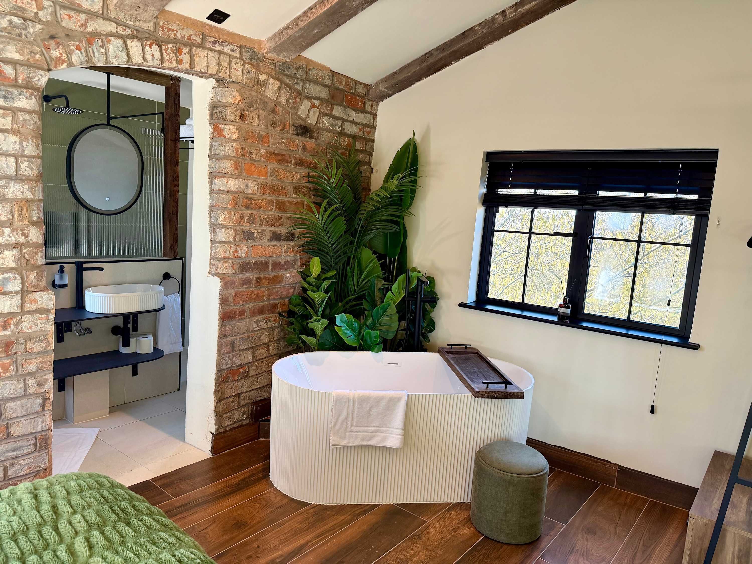 A bathroom with a brick wall, a round mirror, a white sink, a large potted plant, a bathtub, and a window with blinds.