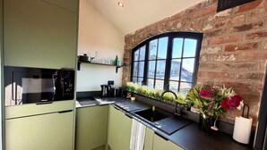 Une cuisine avec des meubles de couleur verte, un plan de travail noir, une fenêtre donnant sur un beau paysage et un mur en briques.