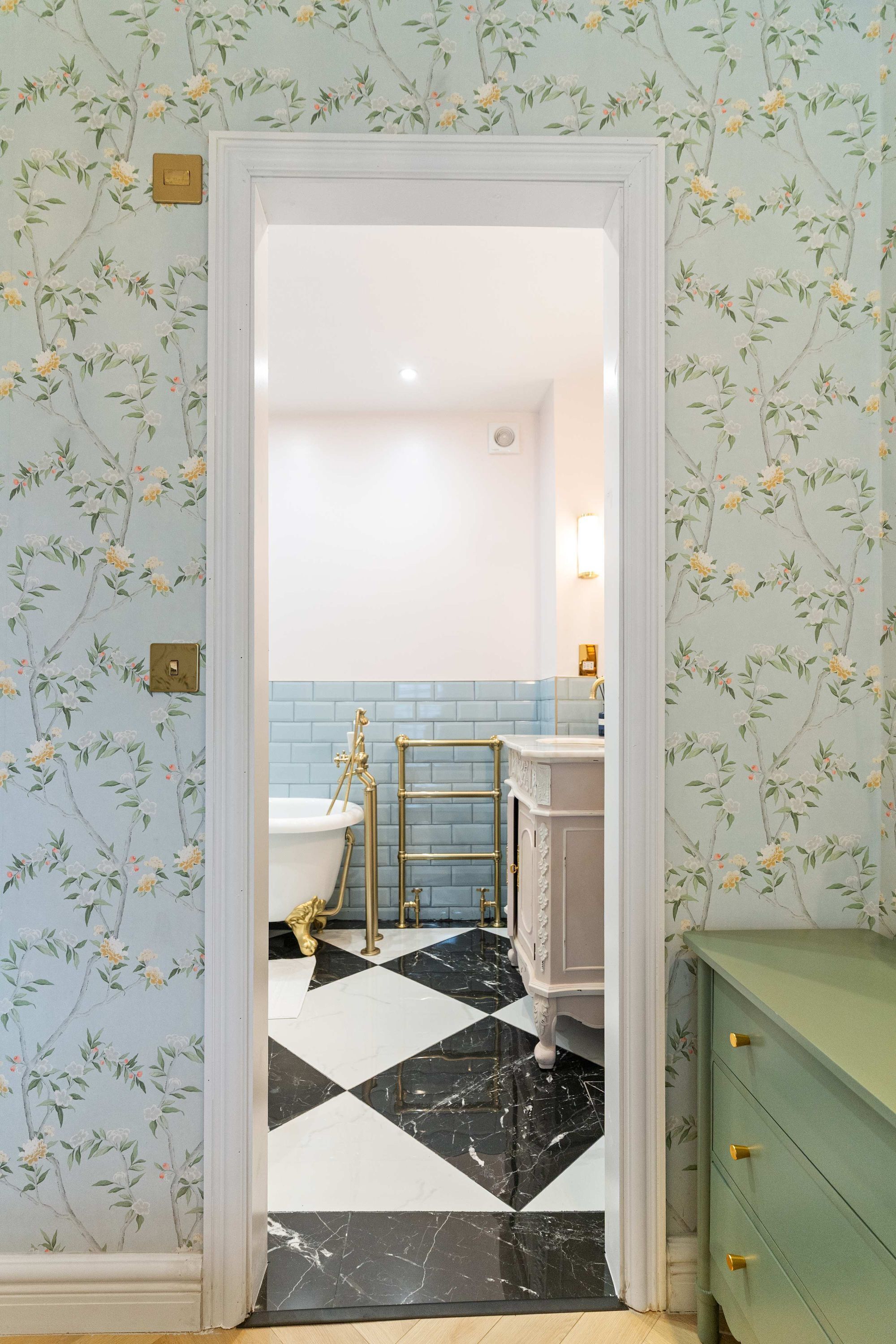 A bathroom with floral wallpaper, a black and white checkered floor, a white bathtub, and a green cabinet.