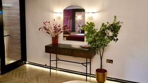 A hallway with a wooden floor, a dark wooden console table with a vase of dried flowers, a potted plant, and a mirror with wall sconces. - Boutique Studio Gold Bath in Jewellery Quarter BHM (Birmingham)