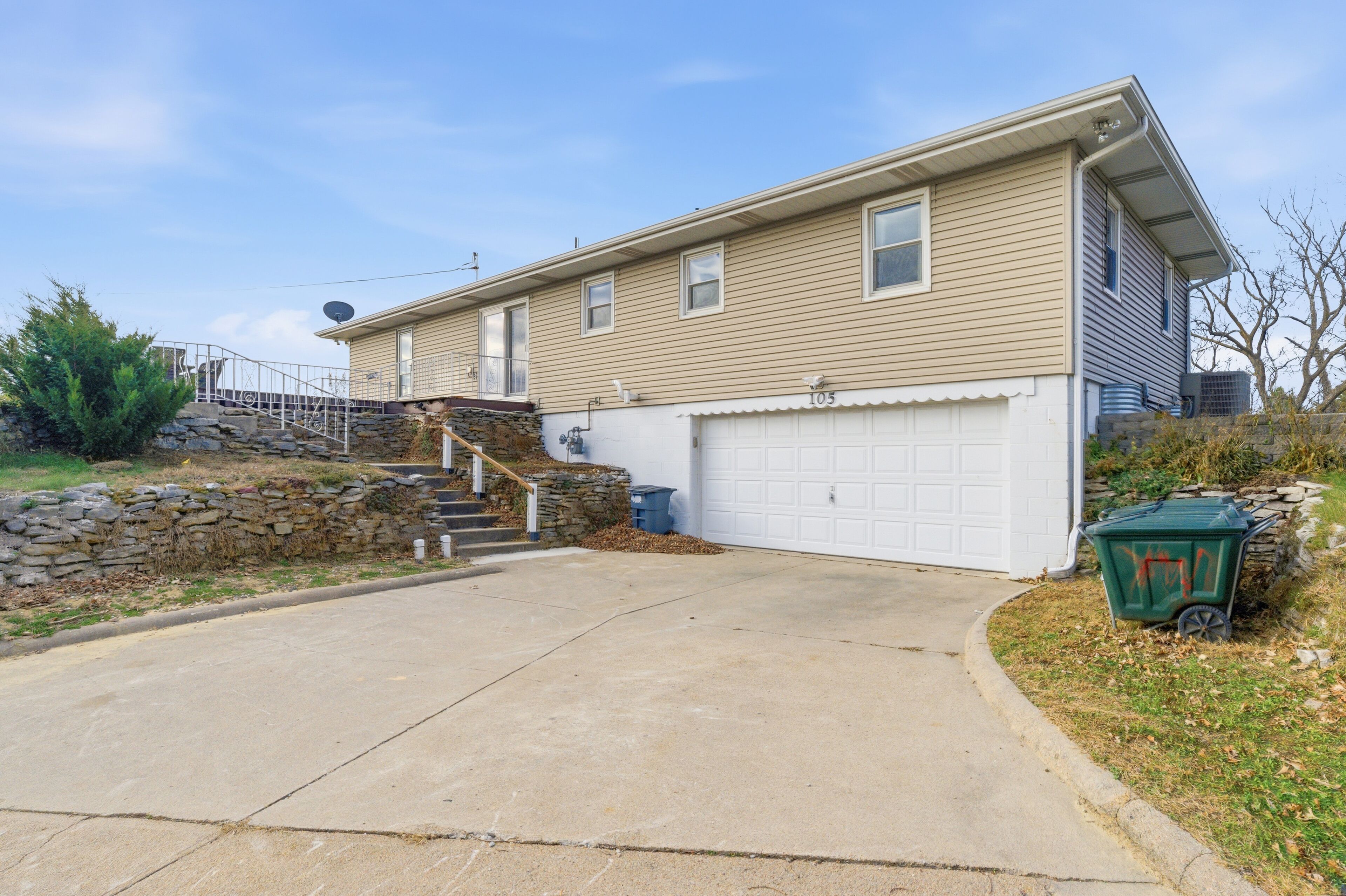A two-story house with a garage, a stone staircase, and a green trash bin.